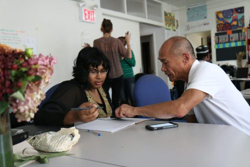 two adults seated at table, one is helping the other with paperwork